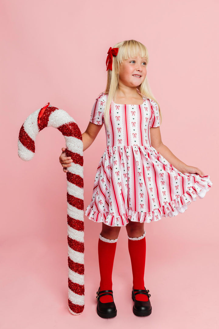 little girl holding giant candy cane in twirl dress with vintage candy cane print on white base paired with red velvet bow and red knee highs with white lace trim