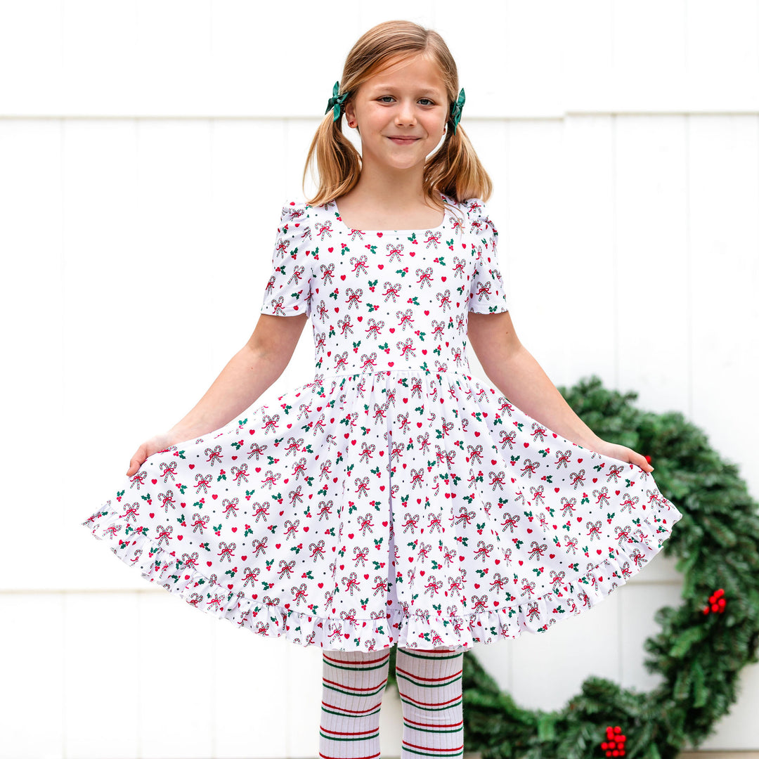 little girl in white Christmas dress with classic red, green and white candy cane and holly print, matching striped tights and green bows