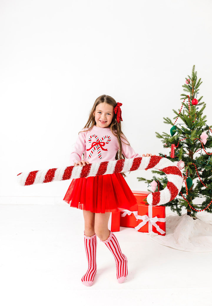 young girl holding oversized candy cane decoration in pink candy cane sweater, red tutu skirt and matching pink lace top knee socks with red vertical stripes