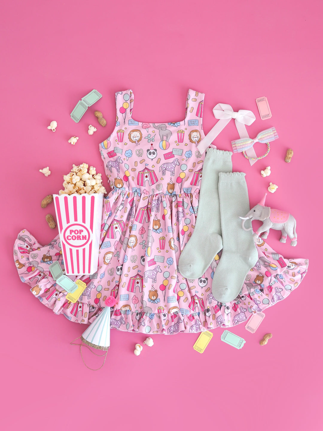 circus themed flatlay of girls' pink sleeveless twirl dress with carnival themed print featuring square neckline and ruffle hem and matching accessories