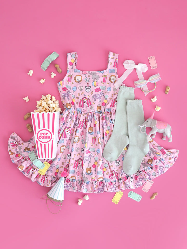 circus themed flatlay of girls' pink sleeveless twirl dress with carnival themed print featuring square neckline and ruffle hem and matching accessories