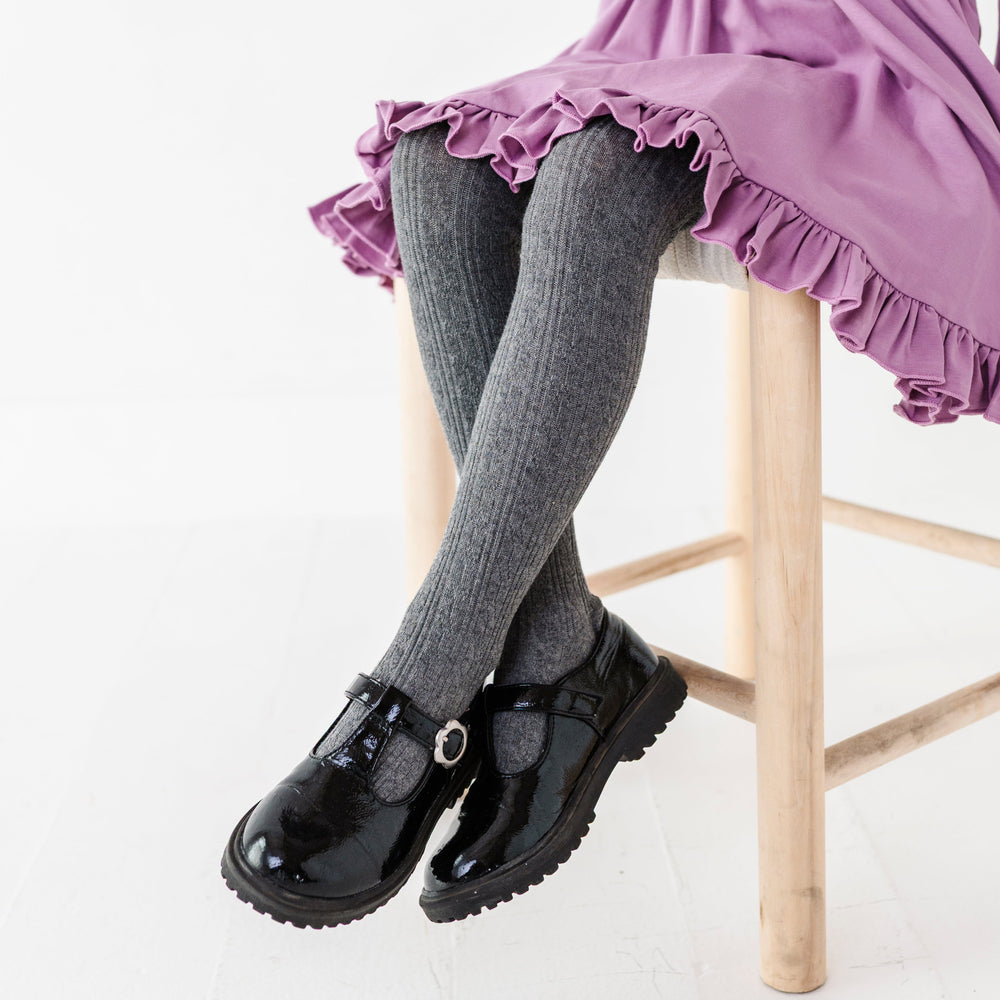 girl sitting on chair with charcoal gray cable knit tights paired with black patent leather mary janes and purple ruffle hem twirl dress