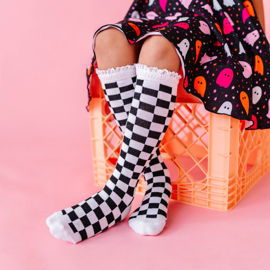little girl sitting on crate in black and white checkered knee socks with lace trim paired with pink and orange ghost print twirl dress for Halloween