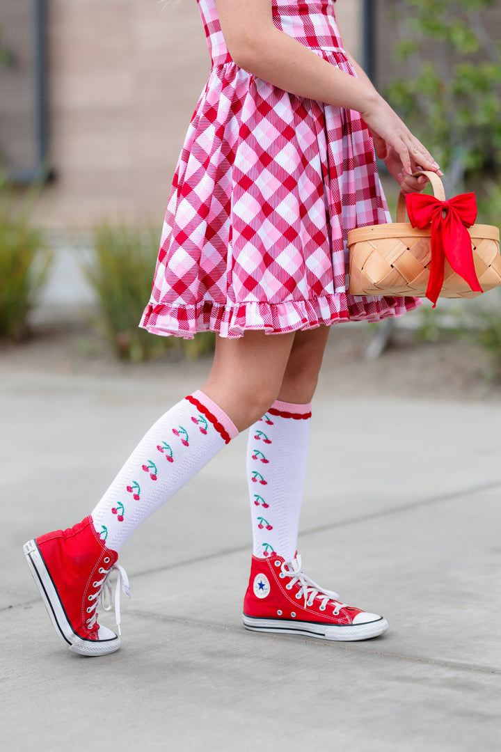 little girl holding a basket of cherries in white knee socks with cherry patter and red high top sneakers paired with red, pink and white plaid twirl dress