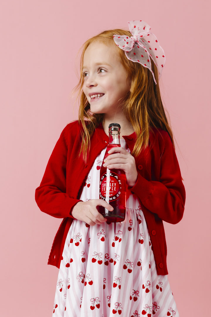 girl holding cherry soda wearing cherry print twirl dress, bright red knit cardigan and matching sheer pink with red dot hair bow