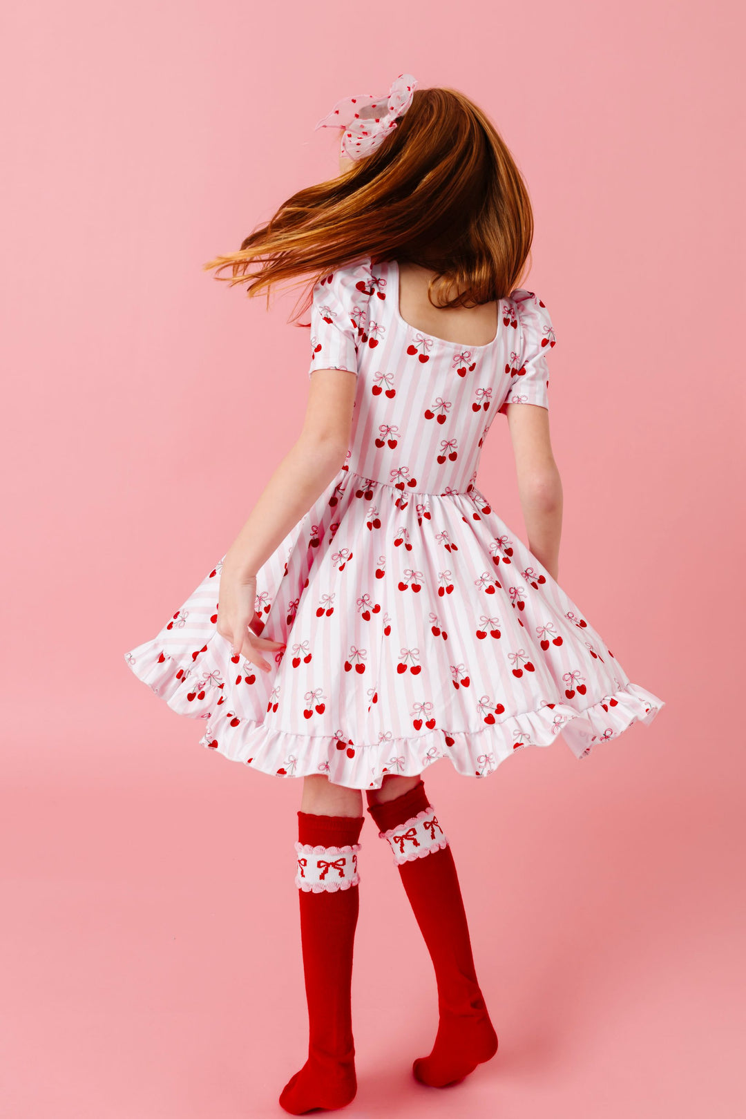 girl twirling in pink and white striped twirl dress with cherry print, sheer pink with red dot hair bow and red scalloped knee socks with bow print