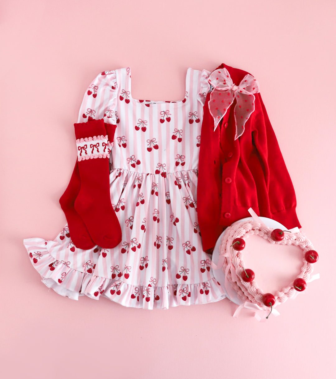girls styled flat lay with cherry themed outfit and heart shaped cake featuring pink and white striped cherry print twirl dress paired with red button front cardigan, matching red socks with bow design and sheer pink bow with red dots