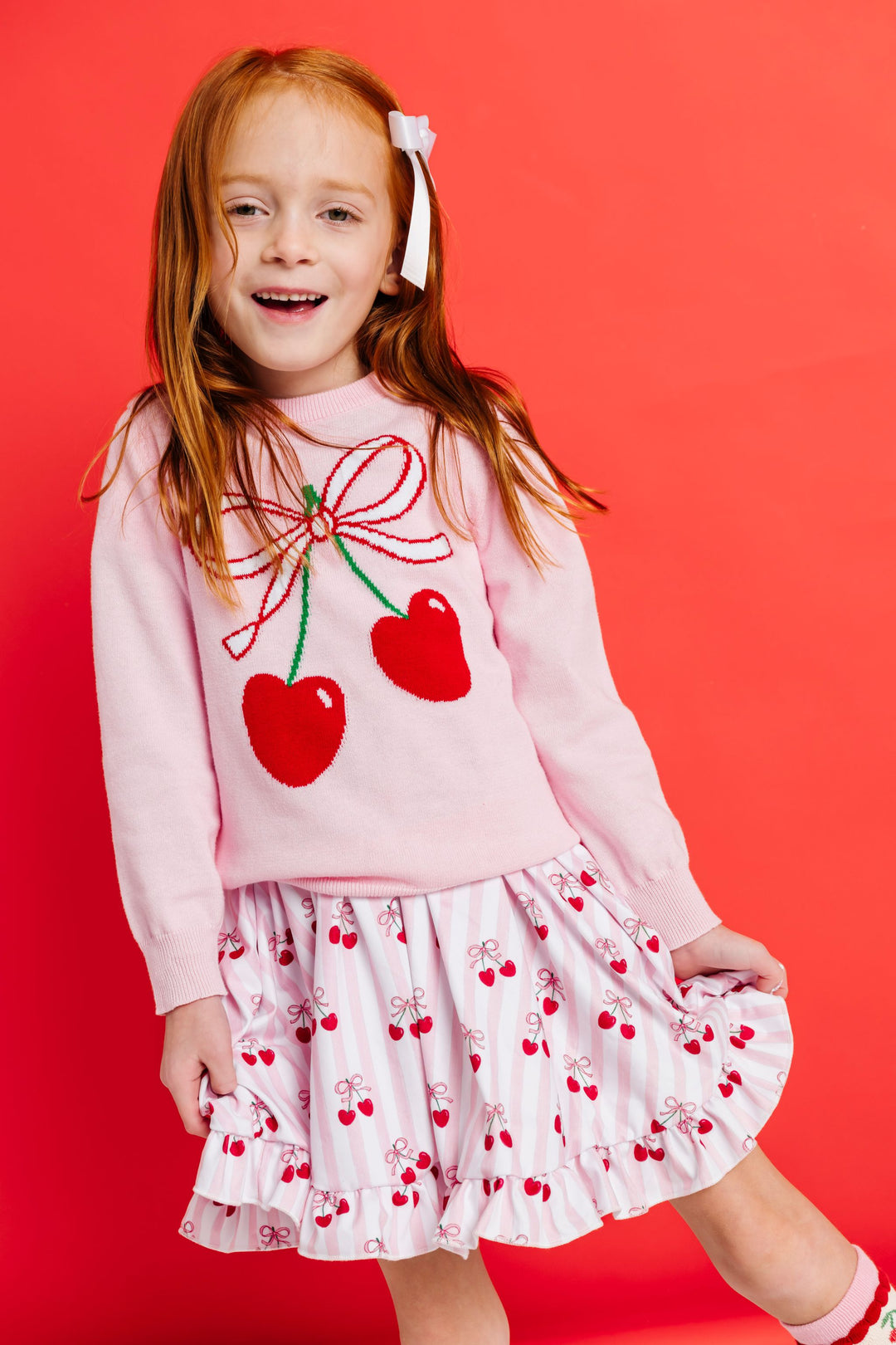 young girl in pink pullover sweater with cherry design paired with matching pink and white striped twirl dress with cherry print