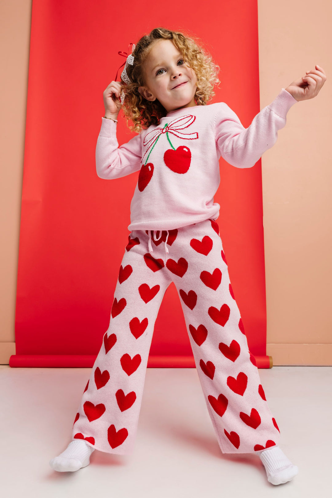 little girl in pink cotton knit sweater set featuring heart shaped cherry design on top and red heart pants for valentines day
