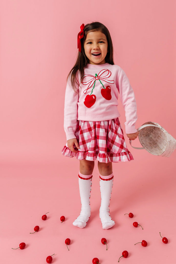 young girl with cherries on the ground around her wearing pink cherry sweater, matching pink and red plaid twirl dress, cherry print knee highs and red satin bow