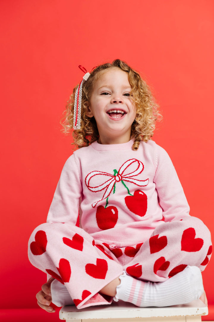 little girl sitting in light pink pullover with cherry and ribbon design paired with pink and red heart sweater pants and matching lace bow