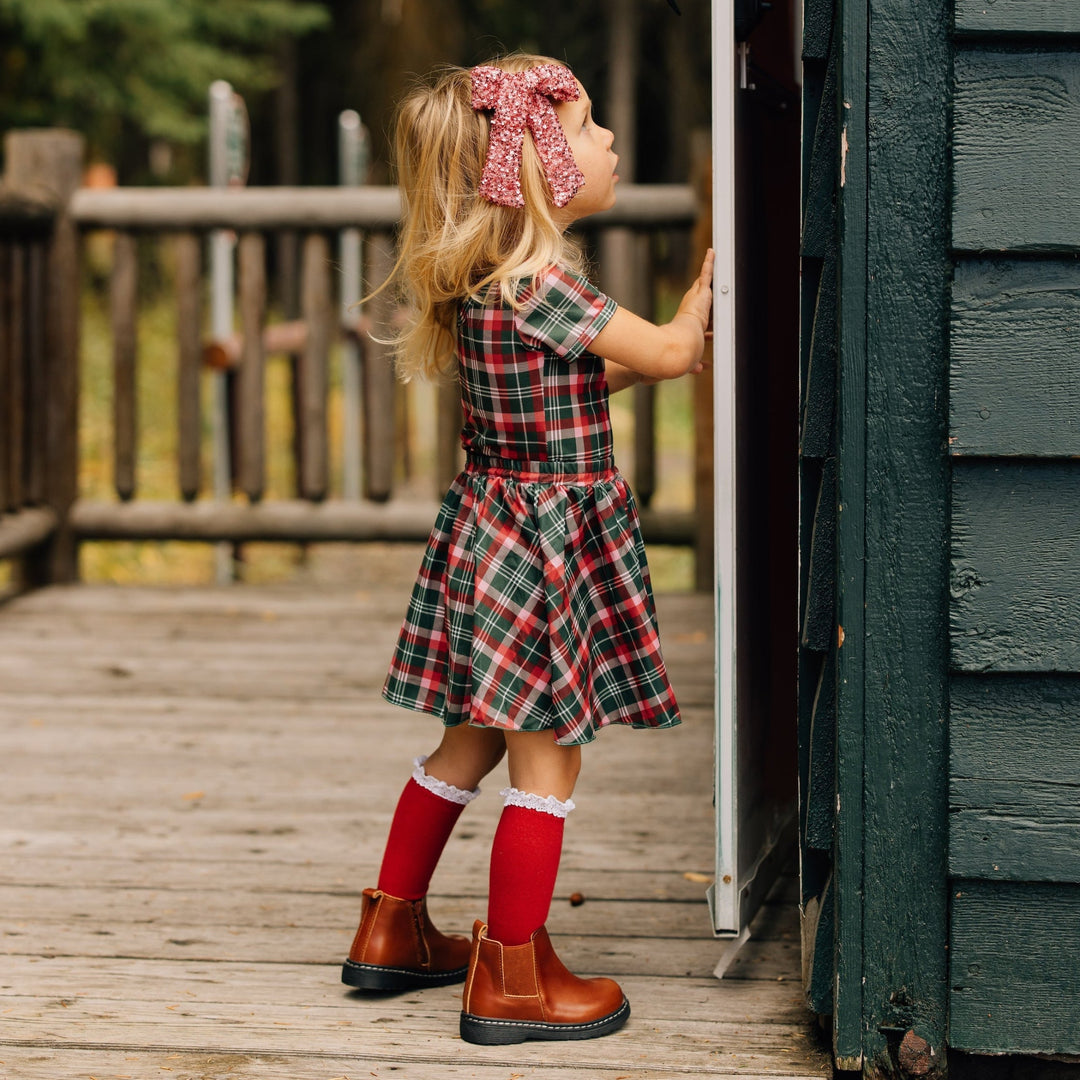 little girl in Christmas plaid top and skirt set paired with red knee-high socks with white lace trim and sparkly pink hair bow