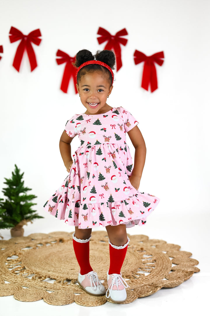 little girl in pink twirly dress with Santa, reindeer and Christmas tree print, red knee highs with white lace trim and red sequin headband