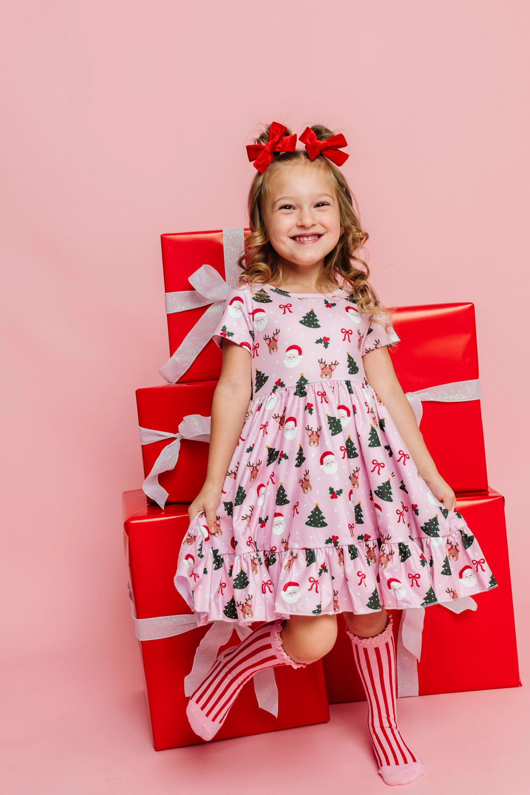 little girl in front of red wrapped Christmas gifts wearing pink dress with Classic Christmas print, red and pink striped knee highs and bright red velvet pigtail bows