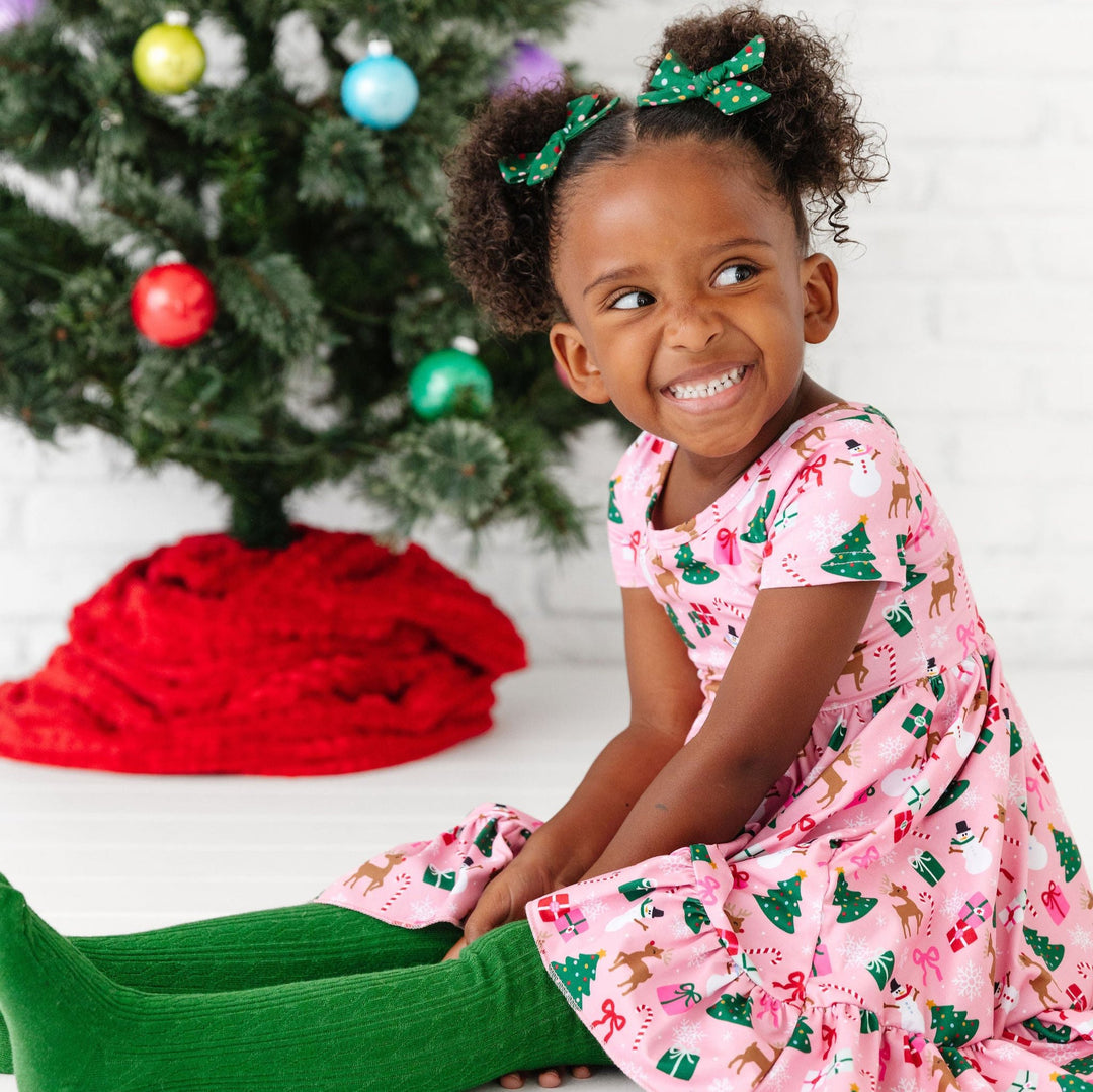 little girl wearing pink christmas print dress with matching hair bows and green cable knit tights