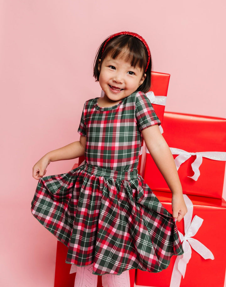 little girl in front of Christmas gifts in red, forest green and pink holiday plaid top and skirt paired with sparkly red headband and pink tights