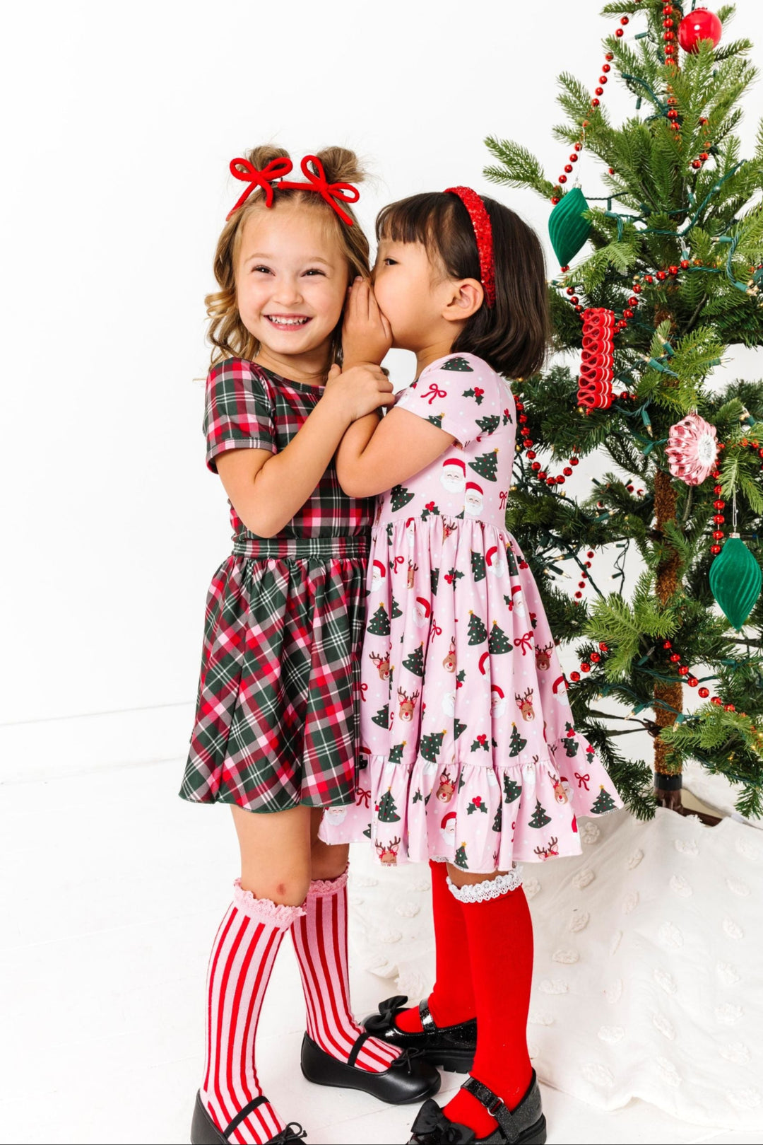 two little girl whispering by Christmas tree in classic Christmas plaid and Santa themed outfits