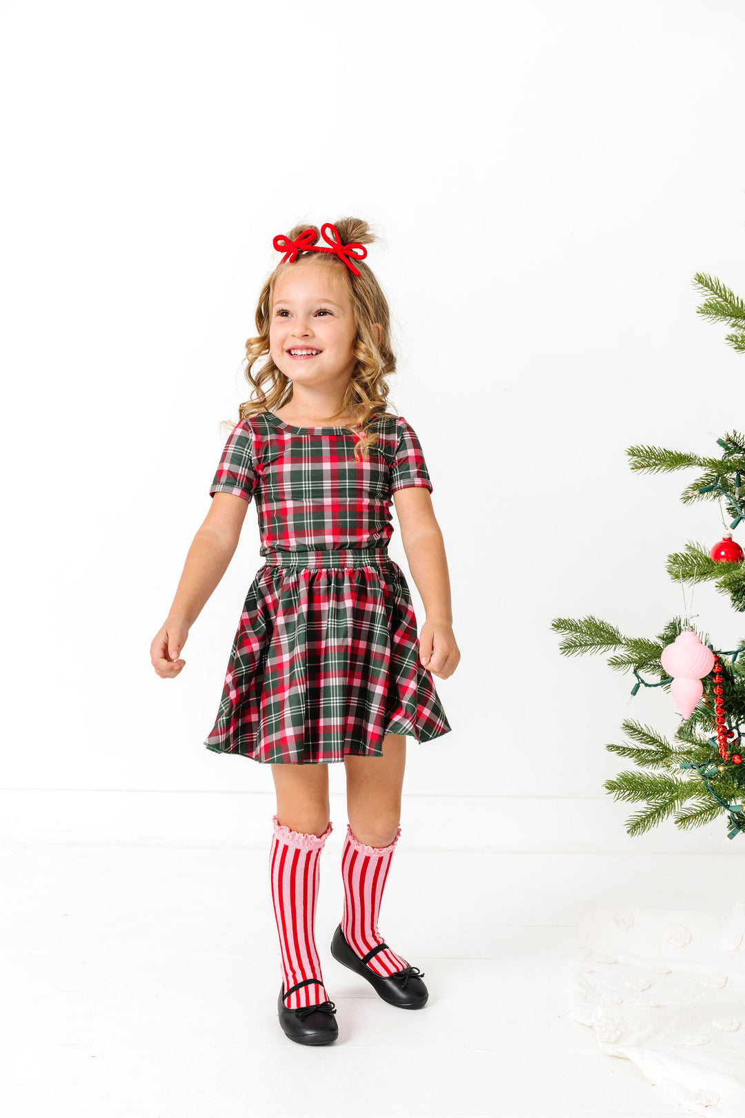 little girl in matching green, red and pink plaid top and skirt paired with red yarn pigtail bows and red and pink striped knee high socks for Christmas