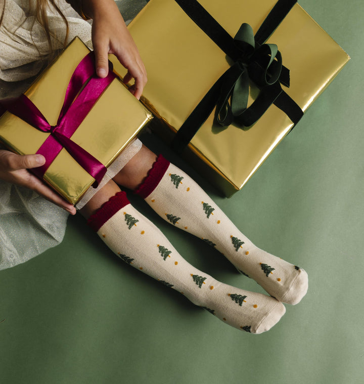 little girl holding gold Christmas gift in sparkly tutu dress and cream knee high socks with crimson scalloped trim and tree pattern