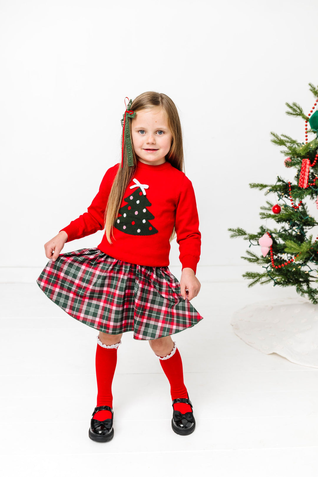 little girl by Christmas tree in red sweater with tree design and 3D white bow topper paired with Christmas plaid skirt and matching accessories