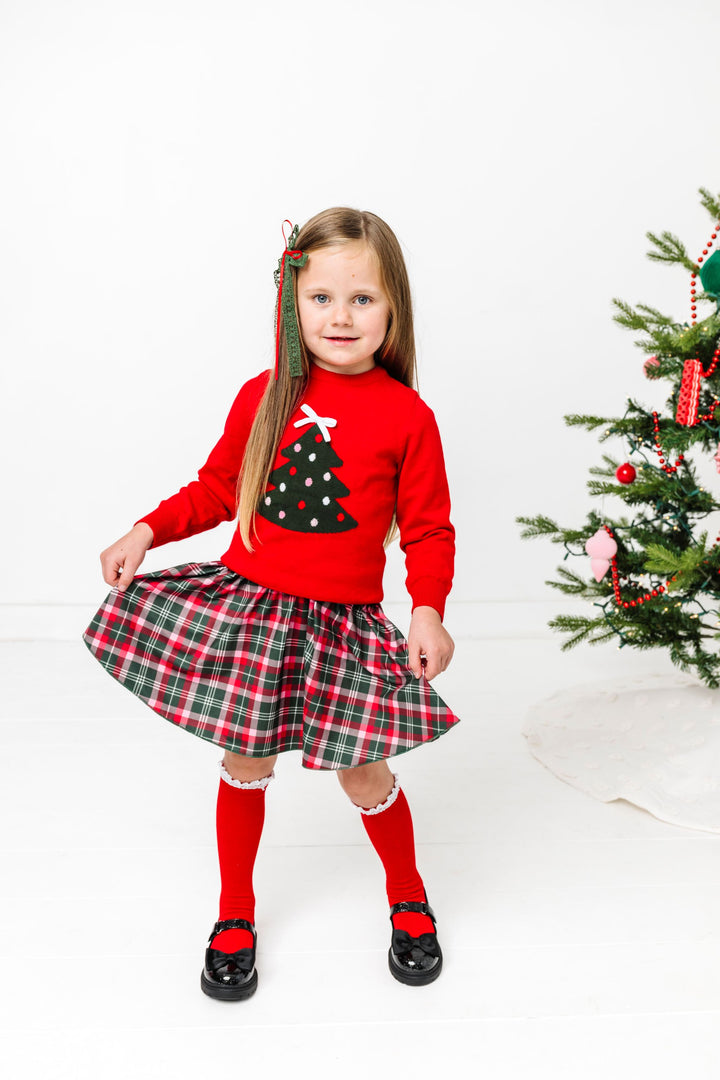 little girl by Christmas tree in red sweater with tree design and 3D white bow topper paired with Christmas plaid skirt and matching accessories