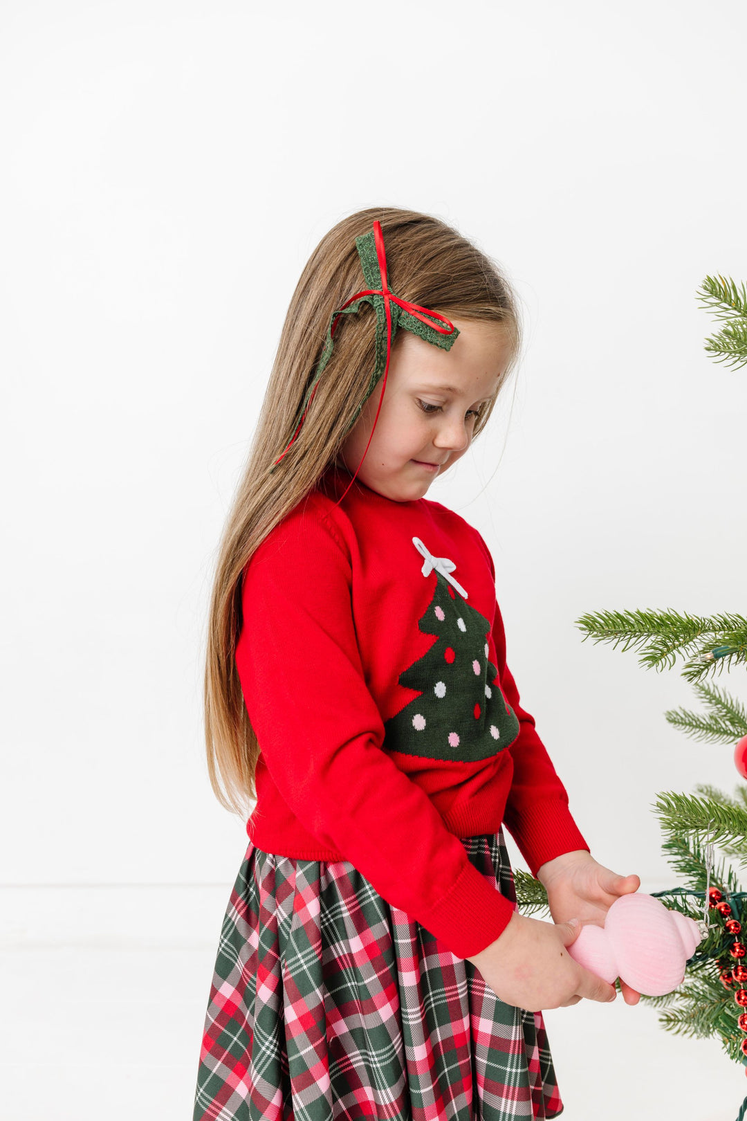 little girl decorating Christmas tree in red sweater with tree design and 3D white bow topper