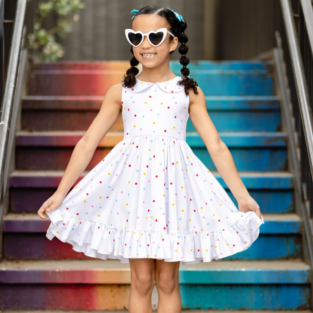 little girl standing in front of bright colored steps wearing white and rainbow polka dot tank style twirl dress