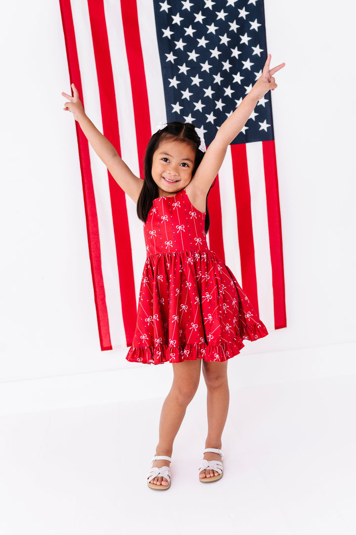 little girl posing in front of American flag in red sleeveless 4th of July twirl dress with white ribbon and blue star print