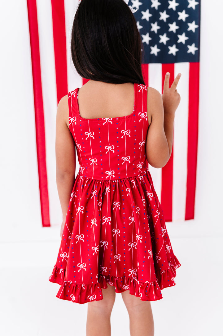 back view of square neck tank twirl dress in red with bow pattern on little girl in front of American flag