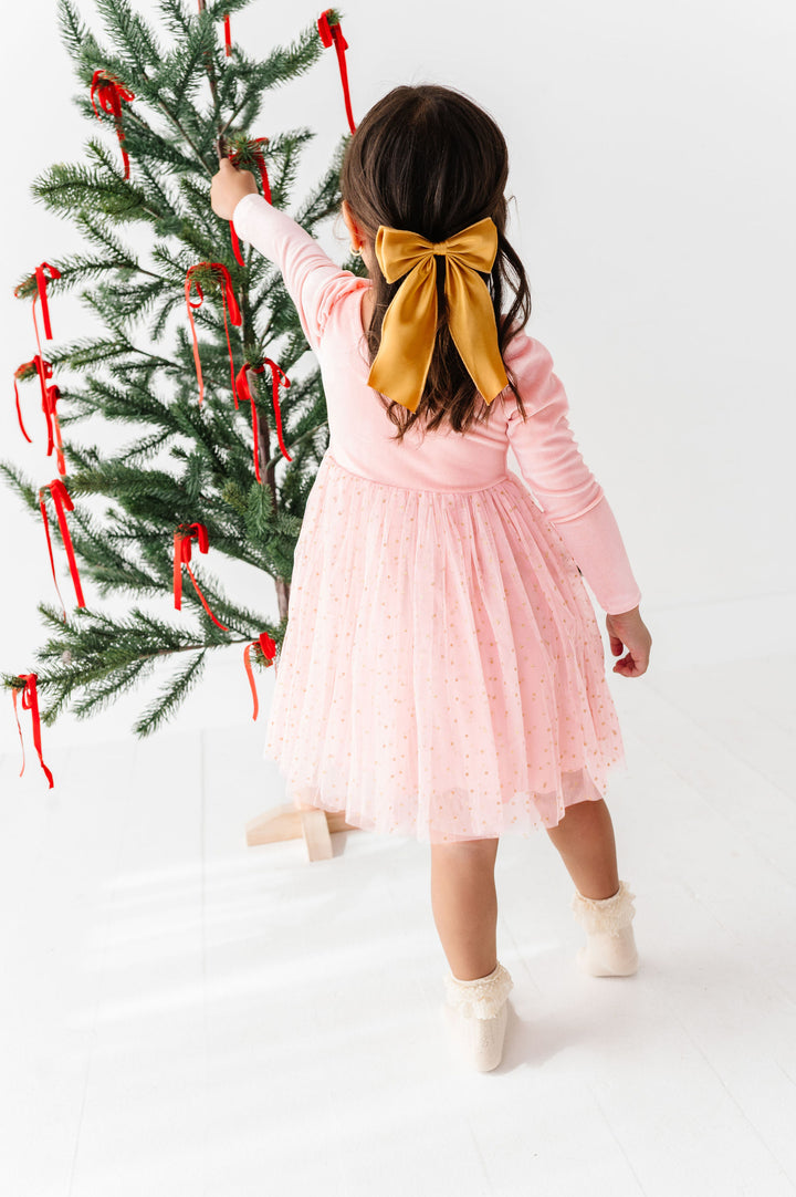 A young girl viewed from behind wearing a light pink long-sleeve tulle dress and a large gold bow in her hair, standing next to a decorated Christmas tree with red ribbons in a bright white room.