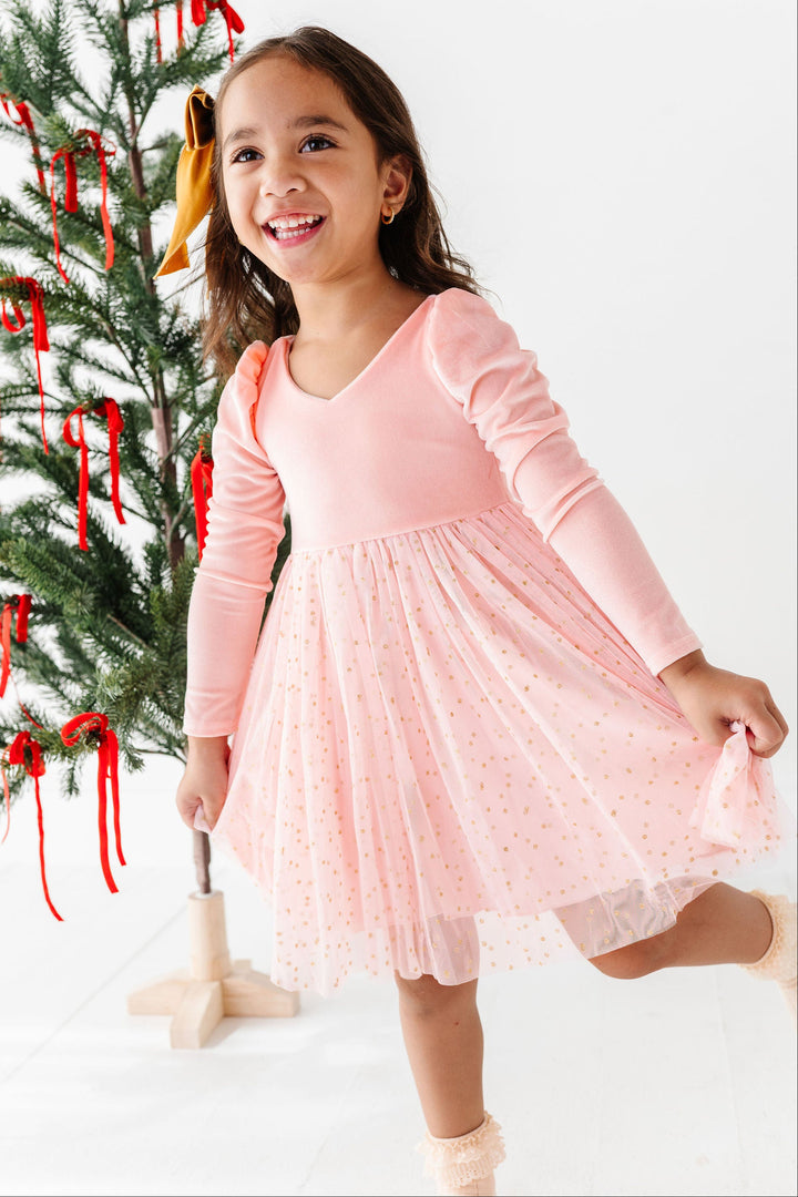 A young girl smiling and twirling in a light pink long-sleeve dress with a polka-dot tulle skirt, standing next to a decorated Christmas tree on a white studio background.