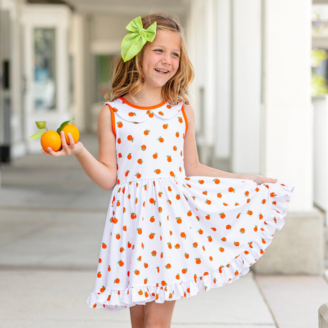 little girl wearing orange print tank top summer dress and holding out ruffle skirt in one hand and holding two oranges in the other