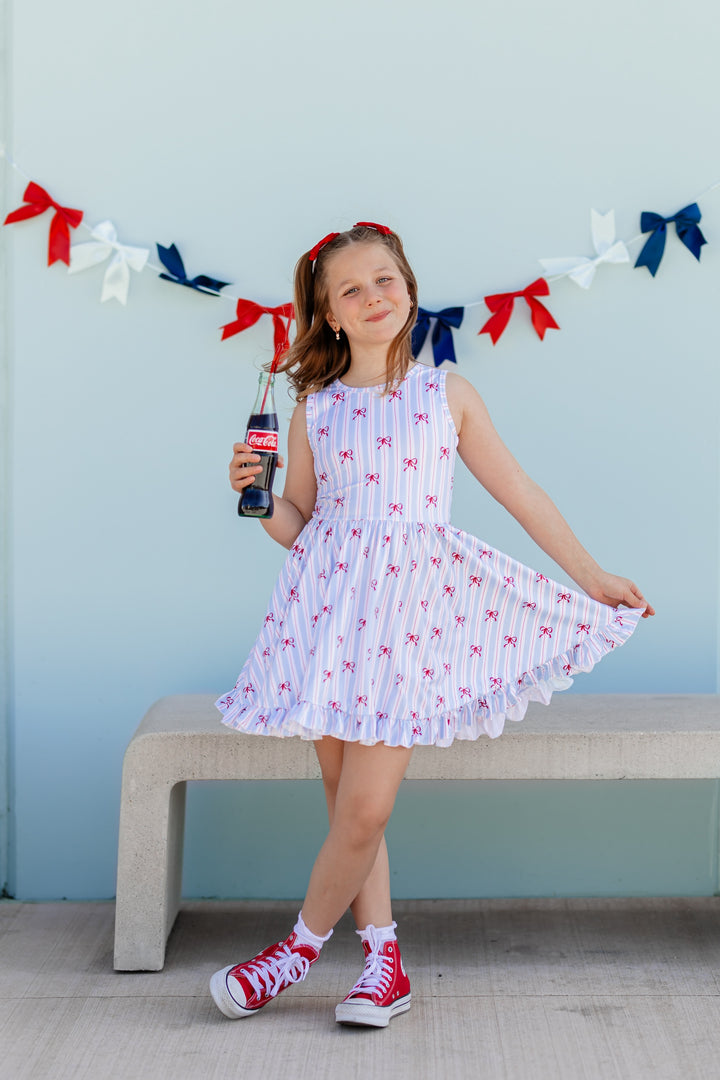 girl in sleeveless twirl dress with white, blue and red stripe with dainty red bow print for 4th of July