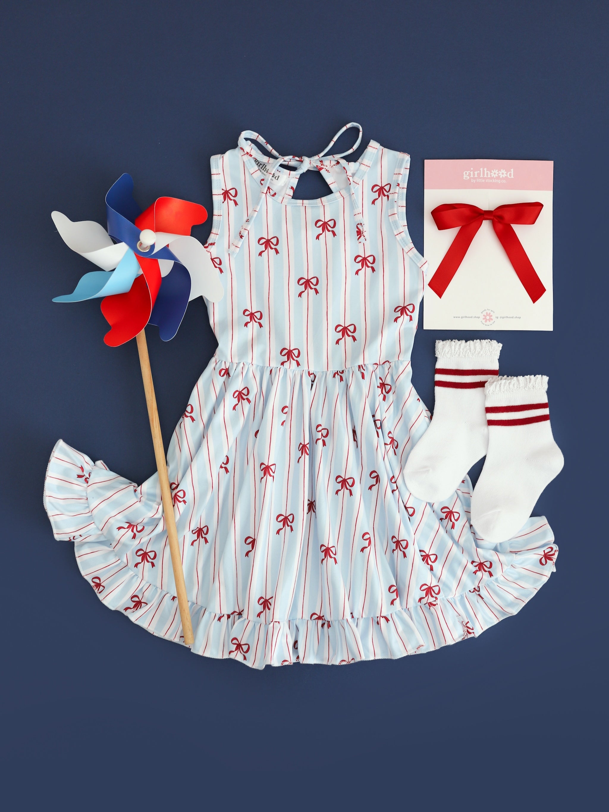flatlay picture of girls sleeveless twirl dress with white, blue and red stripe with dainty red bow print for 4th of July with matching red ribbon bow and striped red midi socks
