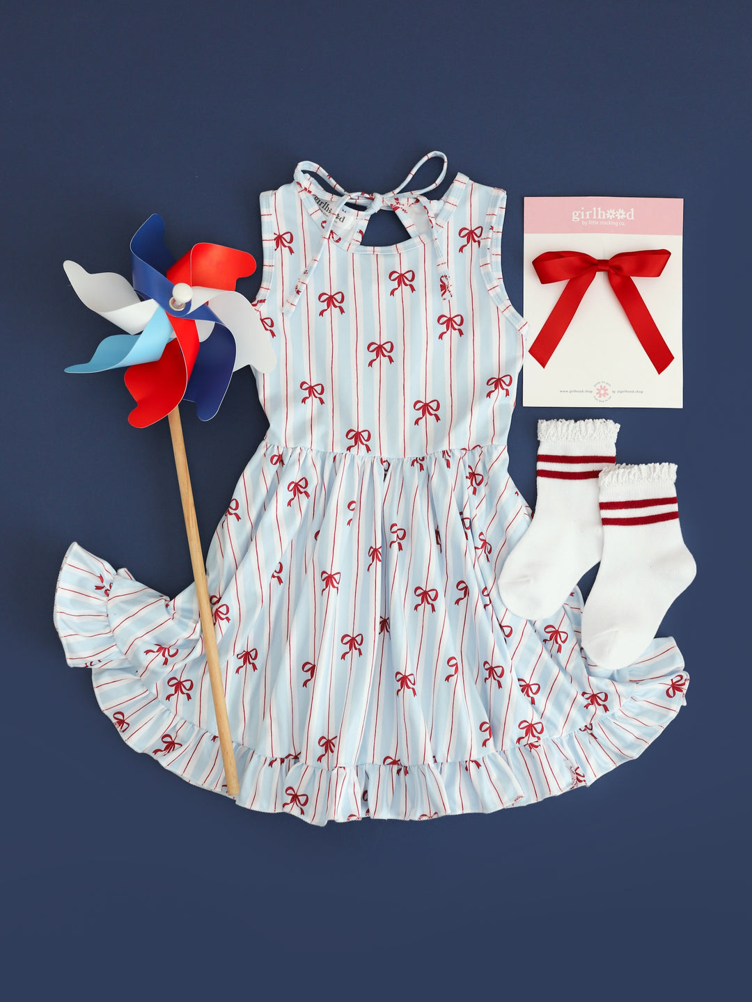 flatlay picture of girls sleeveless twirl dress with white, blue and red stripe with dainty red bow print for 4th of July with matching red ribbon bow and striped red midi socks