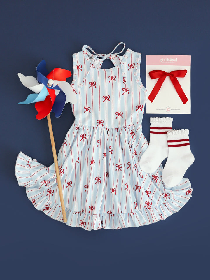 flatlay picture of girls sleeveless twirl dress with white, blue and red stripe with dainty red bow print for 4th of July with matching red ribbon bow and striped red midi socks