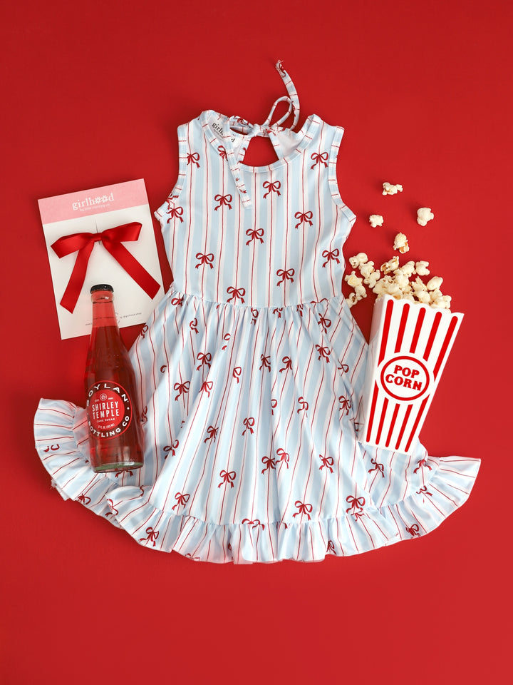 flatlay of girls sleeveless twirl dress with white, blue and red stripe with dainty red bow print for 4th of July and matching red satin ribbon bow