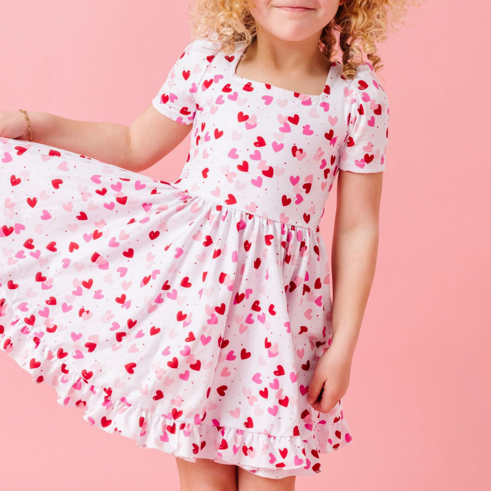 girl in white Valentine's themed dress with scattered hearts print in shades of pink and red paired with pink satin ribbon hair bow and matching pink cable knit knee socks