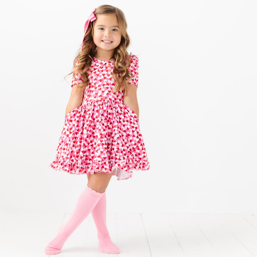 little girl posing in pink, red and white confetti heart print twirl dress and matching pink satin hair bow and knee high socks