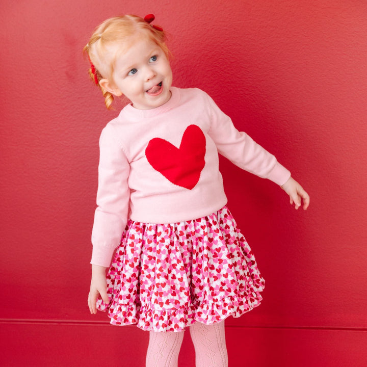 little girl in pink, red and white confetti heart print dress with matching pink and red heart pullover sweater, red bows and light pink pointelle tights