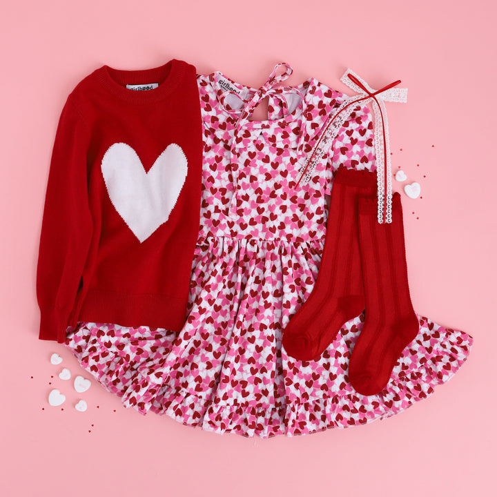 little girl'ss styled Valentine's flat lay outfit with confetti heart print twirl dress, matching red knee highs, red pullover heart sweater and white lace hair bow