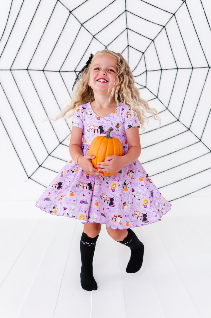 young girl twirling with pumpkin in front of spider web decor in lavender costume kitten print twirl dress and matching black cat socks for Halloween