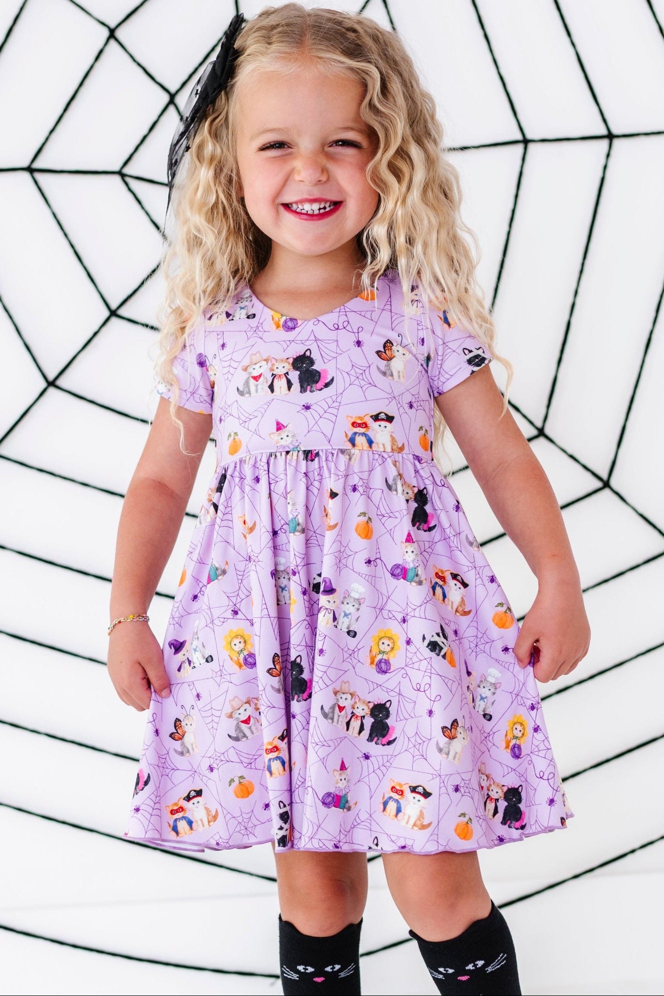 young girl in front of spider web decoration wearing lavender v-neck twirl dress with purple spider web pattern and little kittens dressed up in Halloween costumes