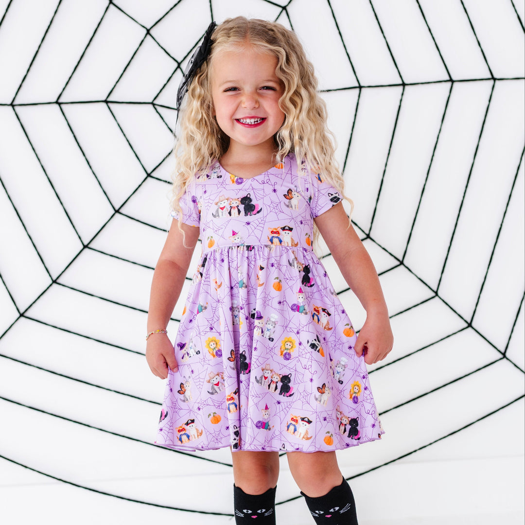young girl in front of spider web decoration wearing lavender v-neck twirl dress with purple spider web pattern and little kittens dressed up in Halloween costumes