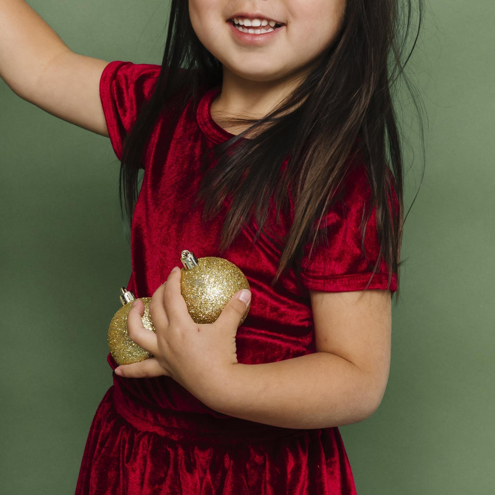 little girl holding gold Christmas ornaments in cranberry velvet top and matching skirt