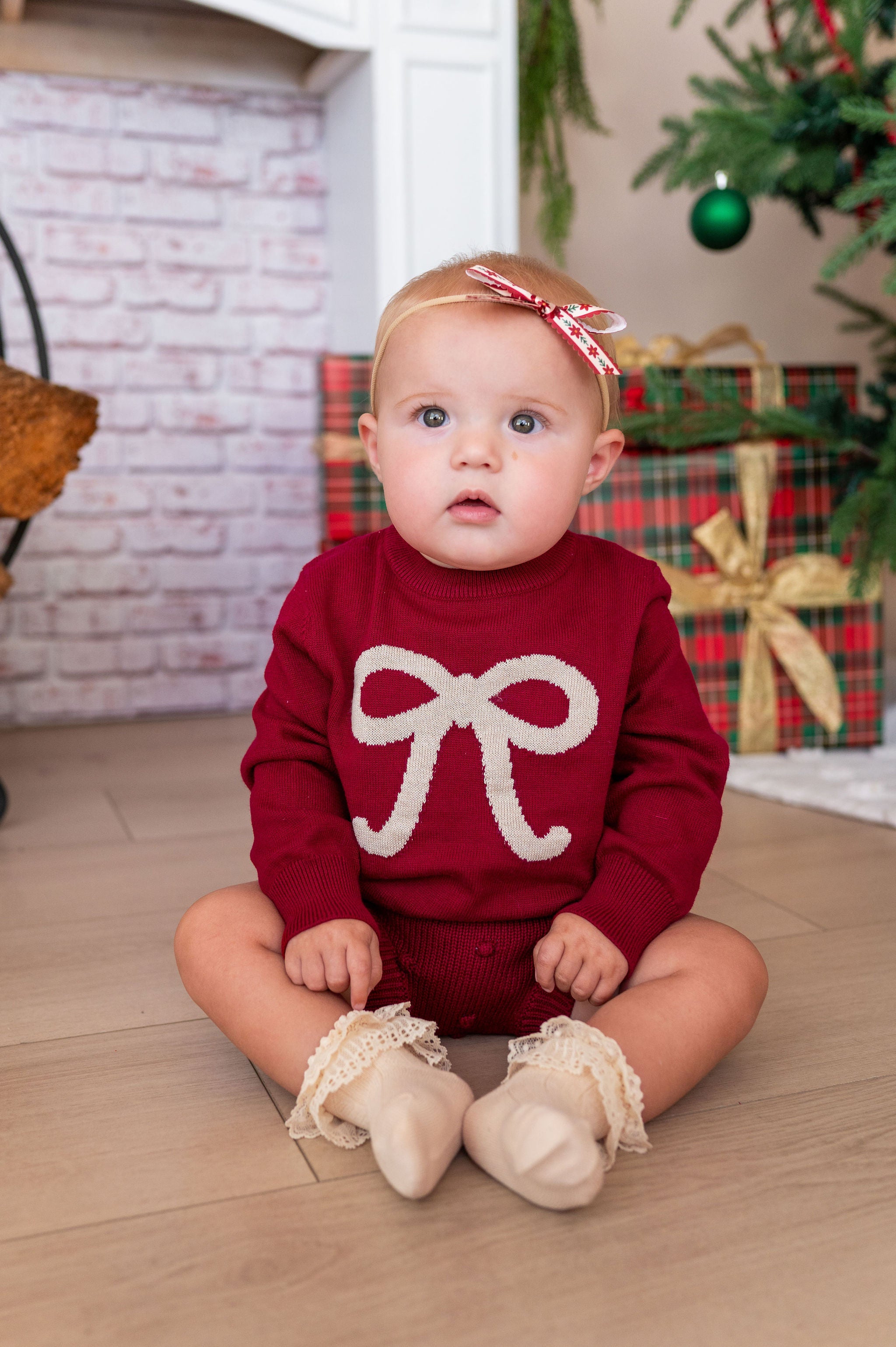 A baby sitting on a wooden floor wearing a festive red knit sweater with a cream bow design, cream lace booties, and a red and white bow headband, with a Christmas tree and wrapped gifts in the background.