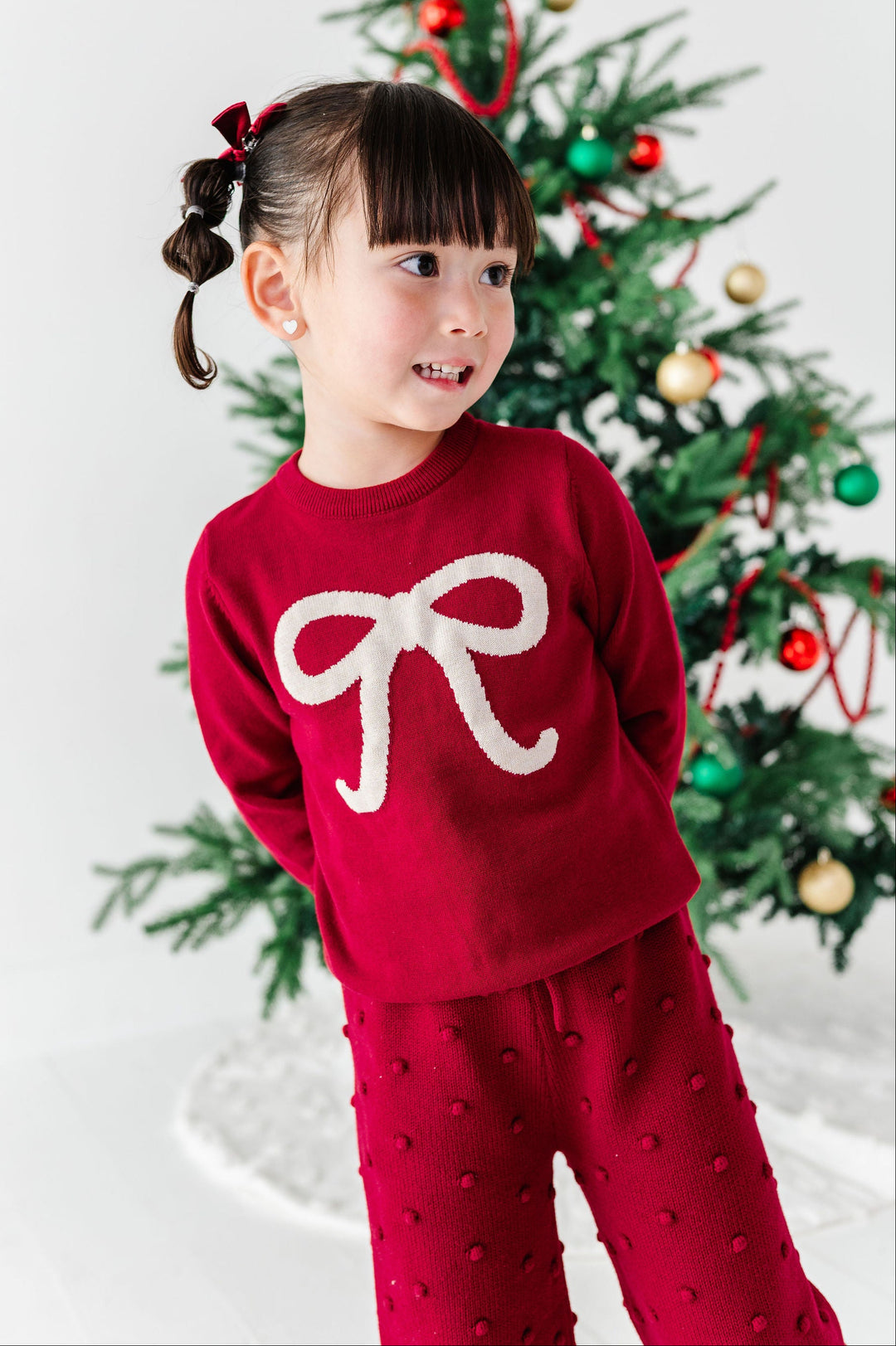 A young girl with pigtails wearing a festive red knit pajama set with a large white bow on the front, standing in front of a decorated Christmas tree.