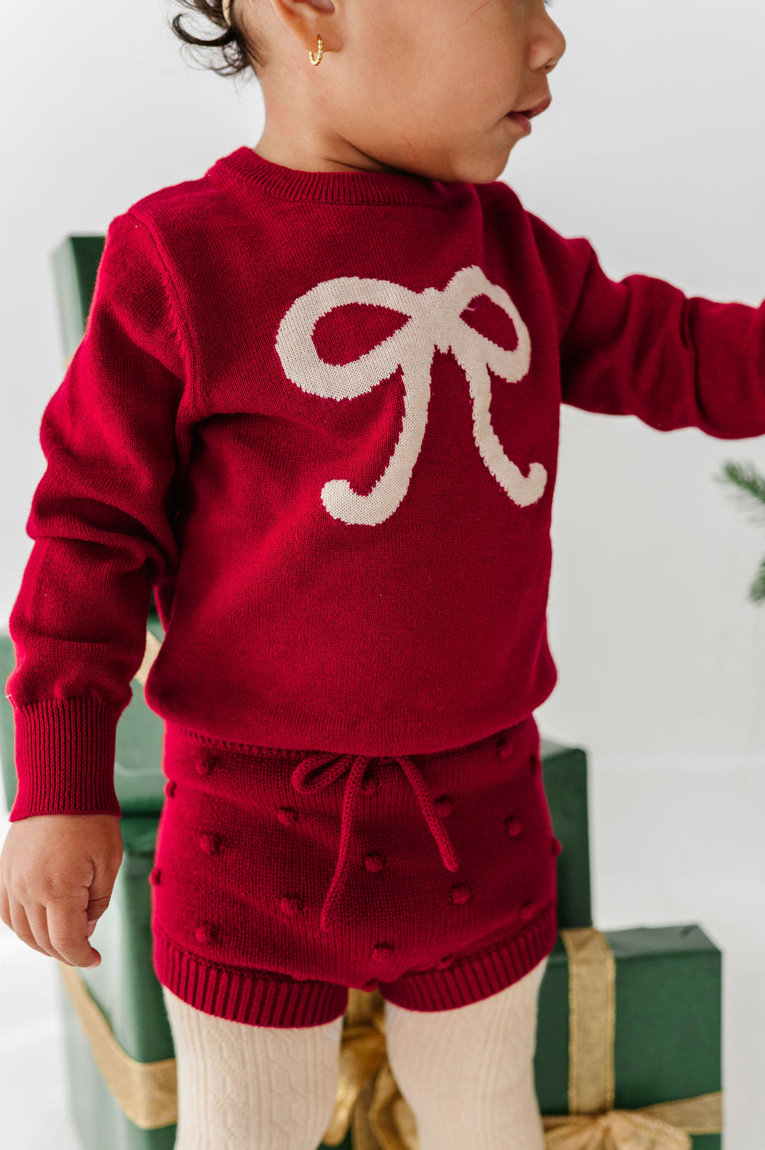 A toddler wearing a festive red knit sweater and matching knit shorts with a cream bow motif on the chest, standing in front of wrapped holiday presents.
