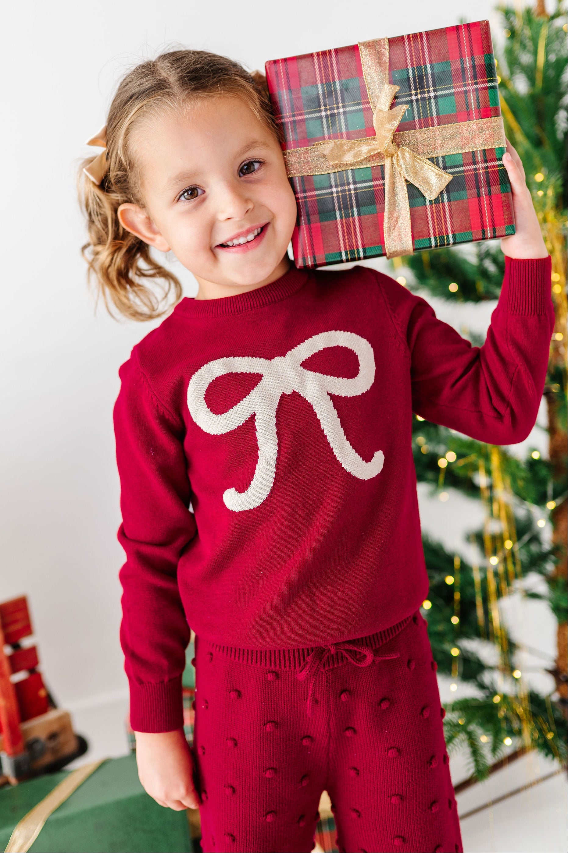 A young girl smiling and holding a wrapped gift box, wearing a festive red long-sleeve sweater with a large white bow motif and matching red patterned pants, set against a decorated Christmas tree background.