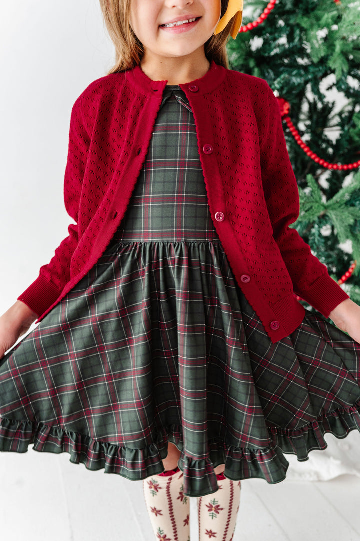 A young girl wearing a festive holiday outfit featuring a dark green and red plaid dress with a matching red button-up cardigan, standing in front of a decorated Christmas tree.